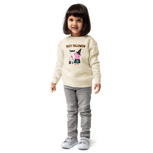 May include: A cream-coloured long-sleeve sweatshirt featuring a graphic of a pink pig character dressed as a witch with a black hat, holding a wand, and a small green creature. The text "HAPPY HALLOWEEN" is printed in brown above the graphic.