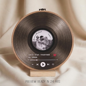 Op de afbeelding: Een rond, donkergrijs decoratief stuk dat lijkt op een vinylplaat, gemonteerd in een houten borduurring. Het midden toont een zwart-wit foto van een kussend stel, met de tekst "Diana & Michael" en "August 10th, 2024". De tekst "LET'S STAY TOGETHER" en "AL GREEN" zijn ook zichtbaar.