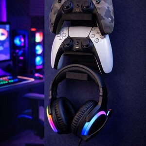 Könnte beinhalten: Eine Wandhalterung für Gaming-Zubehör mit einem Controller im Camouflage-Muster, einem weißen Controller und schwarzen Kopfhörern mit Regenbogen-LED-Lichtern. Die Controller sind über den Kopfhörern gestapelt, alles an einer dunkelblauen Wand montiert.