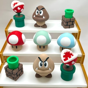 May include: A collection of colorful figurines inspired by a video game. The figurines include red, green, and blue mushrooms with white spots, brown Goombas, and Piranha Plants in green pots. A green pipe is also featured.