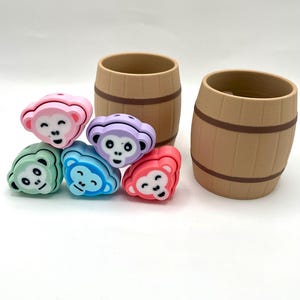 May include: Five colorful monkey-shaped silicone toys with black facial features, stacked on a white surface. Two brown wooden barrels are in the background.