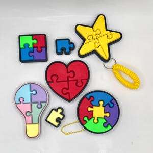 May include: A set of six colorful puzzle pieces shaped like a star, heart, lightbulb, circle, and two squares. Each piece has a different color and is made of soft, flexible material. The pieces are designed to fit together to create a larger puzzle.