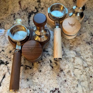 May include: A collection of coffee accessories, including portafilters and tampers. The portafilters have stainless steel baskets and wooden handles. The tampers have wooden handles and stainless steel bases. One tamper has a bird logo.