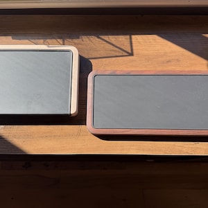 May include: Two rectangular serving trays with dark gray slate centers. The left tray features a light-toned wooden frame, while the right tray has a darker, reddish-brown wooden frame. Both trays have rounded corners and a smooth, flat surface.