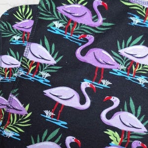 May include: Black fabric with a repeating pattern of lavender flamingos, green tropical leaves, and blue water details. The flamingos have red beaks and legs. The design is suitable for home decor or fashion items.