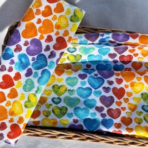 May include: A close-up of several folded cloths with a repeating pattern of colorful hearts in a wicker basket. The hearts are in shades of red, orange, yellow, green, blue, and purple on a white background. The cloths appear to be made of a soft fabric.