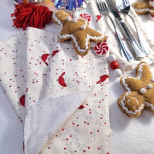 May include: A folded white Christmas-themed cloth napkin with a pattern of red cardinals and berries. The napkin is surrounded by gingerbread man ornaments, silverware, and peppermints, creating a festive holiday scene.