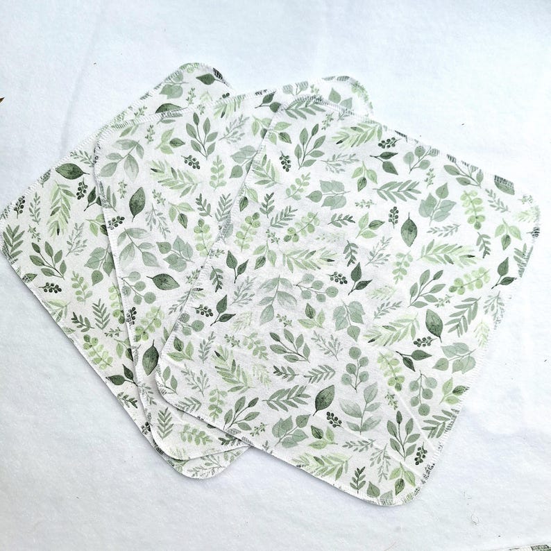 May include: Four white cotton cloths with a green leafy pattern. The cloths are folded and stacked on top of each other.