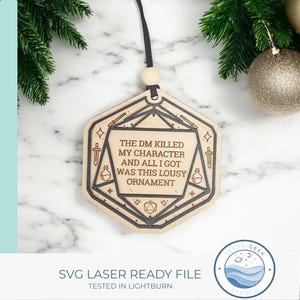 Könnte beinhalten: Weihnachtsornament aus Holz mit einem lasergeschnittenen Design. Das Ornament hat eine hellbraune Farbe und einen schwarzen Rand. Der Text auf dem Ornament lautet: "THE DM KILLED MY CHARACTER AND ALL I GOT WAS THIS LOUSY ORNAMENT". Das Ornament hängt an einem schwarzen Band.