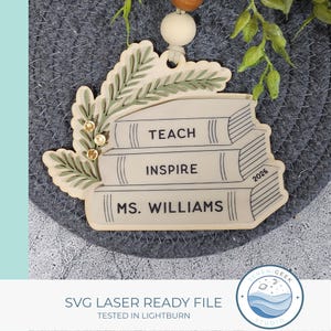 May include: A light-colored wooden ornament shaped like a stack of three books with green leaf accents and three small gold beads. The books are inscribed with the words TEACH, INSPIRE, and MS. WILLIAMS, along with the year 2026 printed on the side.