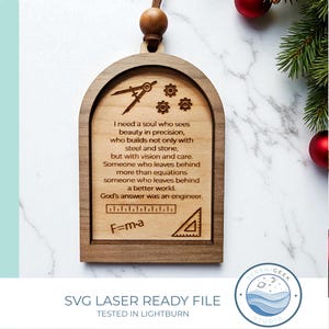 Op de afbeelding: Houten kerstversiering met een gewelfde bovenkant, gegraveerd met een inspirerende quote over ingenieurs, samen met technische symbolen en de formule F=ma. De versiering heeft een houten kraal en een touwtje om op te hangen.