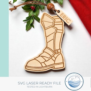 May include: A wooden laser-cut ornament in the shape of a boot, with intricate details. The ornament has a tag that says "ERIC". The ornament is hanging from a leather cord, with green leaves and red berries in the background. The text "SVG LASER READY FILE" is at the bottom.