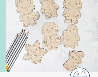 May include: Seven light-colored wooden cutouts featuring engraved outlines of a werewolf, mummy, Frankenstein monster, vampire, ghost with a pumpkin, bat, and cat wearing a hat, accompanied by five colored pencils and the text SVG LASER READY FILE.