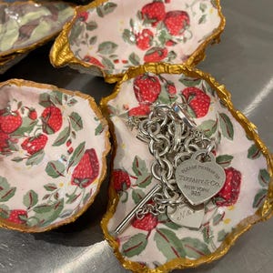 Puede incluir: Cuatro platos de cerámica blanca en forma de concha de ostra con ribete dorado y un patrón de fresas. Los platos están llenos de un collar de cadena de plata y un dije de corazón de plata con el texto "PLEASE RETURN TO TIFFANY & CO. NEW YORK 925" y un dije de corazón más pequeño con las iniciales "N &"