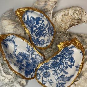 May include: Three decorative oyster shells with a white and blue floral pattern. The edges are trimmed with gold paint. The shells are arranged on a bed of other oyster shells.