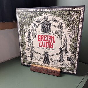 Op de afbeelding: Een vierkante vinyl platenhoes getiteld Green Lung Woodland Rites met een zwart-witte illustratie van figuren in een cirkel rond een centraal gehoornd wezen, met rode tekst, rechtop gehouden door een rechthoekig stuk natuurlijk hout.