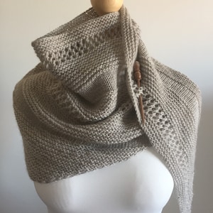 May include: A beige knitted shawl with a delicate lace pattern draped over a white mannequin.