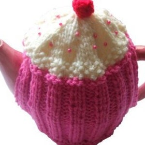 May include: A pink and white knitted teapot cozy shaped like a cupcake with white pearl-like beads and a red pom-pom on top.
