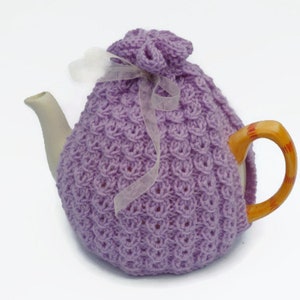 Könnte beinhalten: Ein lila gestrickter Teekannenwärmer mit einem weißen Band. Der Wärmer bedeckt eine Teekanne mit einem weißen Korpus und einem braunen Henkel.