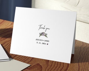 Puede incluir: Una tarjeta de agradecimiento blanca con un diseño floral y el texto "Thank you Jessica + Jared 5.31.2024". La tarjeta está sobre una mesa de madera con un tazón de vidrio de caramelos y un sobre beige.