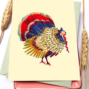 May include: A greeting card featuring a colorful illustration of a turkey. The turkey has a vibrant red, blue, and yellow tail feathers. The card is on a light yellow background, with a green envelope and a pink tray underneath.