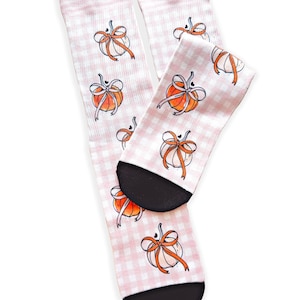 Puede incluir: Un par de calcetines con un patrón de cuadros vichy en rosa claro y blanco. Los calcetines están decorados con un estampado repetitivo de calabazas naranjas, cada una atada con una cinta naranja fina. Las punteras y talones son negros.