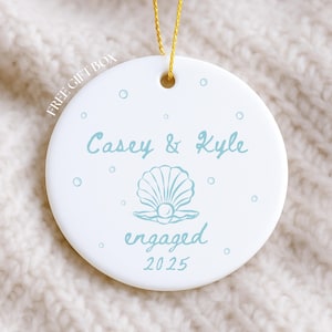 May include: White ceramic ornament with a gold string for hanging. The ornament features the names "Casey & Kyle" in light blue script, along with a seashell design and the word "engaged" and the year "2025".