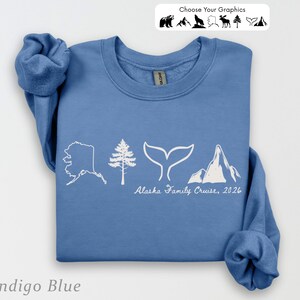 May include: An indigo blue crewneck sweatshirt featuring a white graphic print of the state of Alaska, a pine tree, a whale tail, and a mountain range. Below the graphics, the text reads Alaska Family Cruise, 2026 in a white script font.