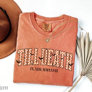 May include: A burnt orange t-shirt featuring the text TILL DEATH in a white and orange checkered pattern with small black bat silhouettes. Below the text, the Roman numerals IX.XIII.MMXXVII are printed in black. The shirt has a crew neckline.