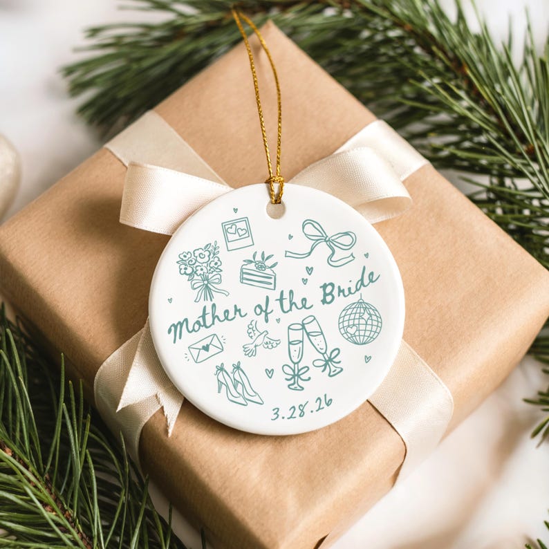 May include: A white ceramic ornament with a gold string, featuring the text "Mother of the Bride" and wedding-themed illustrations in teal. The ornament is placed on a wrapped gift with a cream ribbon, surrounded by greenery.