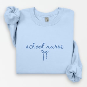 May include: A light blue sweatshirt with the words "school nurse" in a script font, and a bow graphic. The sweatshirt is folded with the sleeves rolled up.