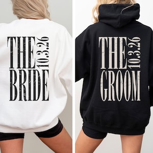 May include: A white sweatshirt and a black hooded sweatshirt, both featuring back prints with distressed lettering. The white sweatshirt displays THE BRIDE and 10.3.26 in black, while the black hoodie features THE GROOM and 10.3.26 in white.