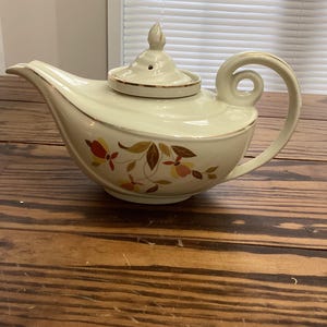 May include: Cream-colored vintage teapot with a curved handle and spout. The lid has a small finial and a gold rim. Floral and leaf designs in red, orange, and brown adorn the side. The teapot sits on a wooden surface.