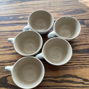 May include: Five white ceramic mugs with a light brown interior. The mugs are arranged in a circle on a wooden surface.