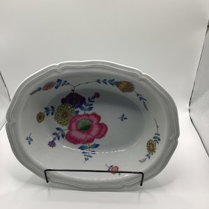 May include: White oval serving dish with a scalloped edge. The dish features a floral design with pink, purple, yellow, and blue flowers. The dish is supported by a black metal stand. A decorative piece for serving.