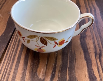 May include: Cream-colored teacup with a gold rim and handle. The cup features a floral pattern with red, yellow, and brown leaves. The teacup is set on a wooden surface.