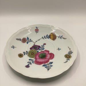 May include: White porcelain saucer with a scalloped edge, decorated with a floral design. The flowers are in shades of pink, yellow, purple, and green, with blue leaves and stems. A delicate piece for serving.