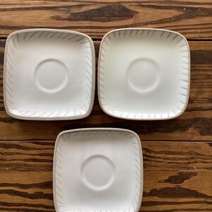 May include: Three white ceramic square saucers with a raised rim and a subtle gray line design. The saucers are perfect for serving coffee or tea.