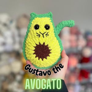 May include: A crocheted avocado-shaped plush toy with a cat face, green top, yellow body, and brown seed. The toy is being held by a hand and the text "Gustavo the AVOGATO" is below the toy.