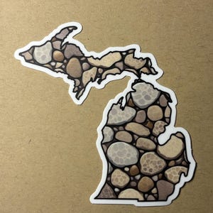 May include: A sticker in the shape of the state of Michigan, featuring a mosaic design of various-sized stones in shades of brown, tan, and gray. The sticker has a white border and is set against a tan background.