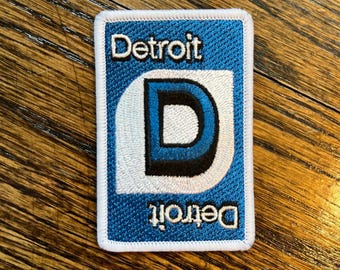May include: A blue and white embroidered patch with the word "Detroit" and a stylized "D" in the center.