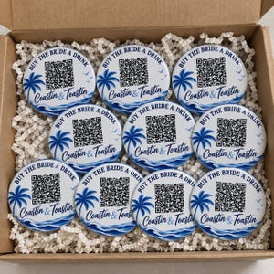 May include: Nine round white buttons featuring a blue palm tree and ocean wave graphic, a black QR code, and the text "BUY THE BRIDE A DRINK" and "Coastin & Toastin" printed in blue. The buttons are arranged in a grid inside a cardboard box.