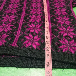 May include: Black and fuchsia-colored knitted sweater with a snowflake pattern. The sweater has a ribbed hem and cuffs. A pink measuring tape is placed vertically on the sweater.