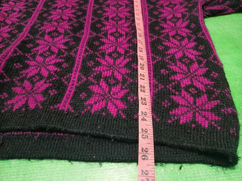 May include: Black and fuchsia-colored knitted sweater with a snowflake pattern. The sweater has a ribbed hem and cuffs. A pink measuring tape is placed vertically on the sweater.