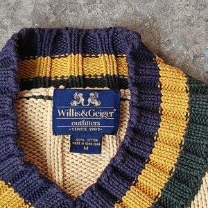 May include: Close-up of a V-neck sweater with a ribbed knit design. The sweater features a navy blue collar and stripes in gold and dark green. A blue label reads "Willis & Geiger outfitters SINCE 1902".
