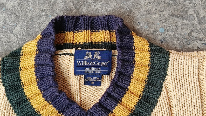 May include: Close-up of a V-neck sweater with a ribbed knit design. The sweater features a navy blue collar and stripes in gold and dark green. A blue label reads "Willis & Geiger outfitters SINCE 1902".