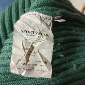 May include: Close-up of a green knit sweater with a tag. The tag reads "SPORTSWEAR COMPANY S.P.A." and includes details about the wool composition and origin. The sweater is made of 100% wool.