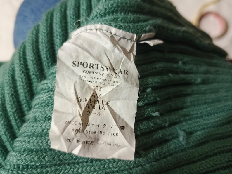 May include: Close-up of a green knit sweater with a tag. The tag reads "SPORTSWEAR COMPANY S.P.A." and includes details about the wool composition and origin. The sweater is made of 100% wool.
