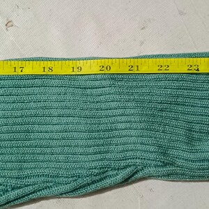 May include: Close-up of a section of a teal knitted fabric. A yellow measuring tape is laid across the fabric, showing a width of approximately 8 inches. The fabric has a ribbed texture.