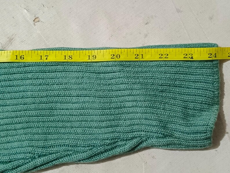 May include: Close-up of a section of a teal knitted fabric. A yellow measuring tape is laid across the fabric, showing a width of approximately 8 inches. The fabric has a ribbed texture.